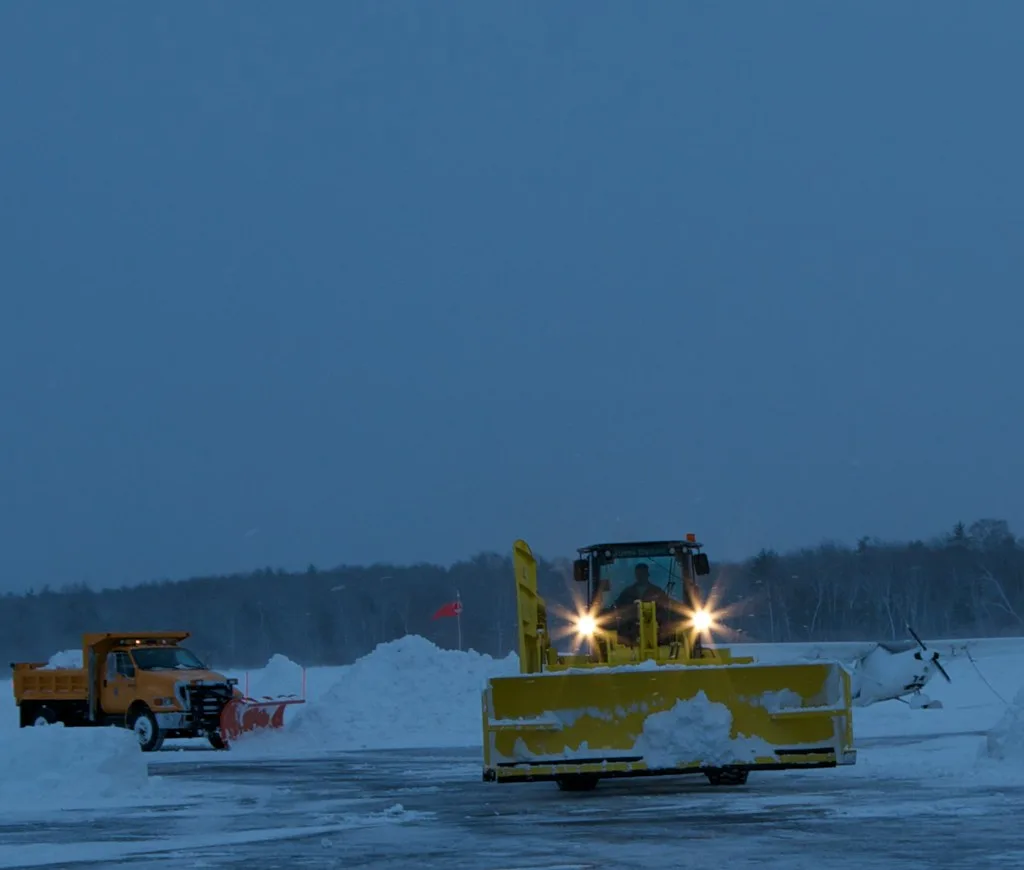 A plow truck and a loader with a pusher box clearing a large paved lot at dusk during a snowstorm